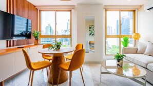A modern living room with a sofa, a dining table with chairs, a TV, and large windows with a city view. - w Charming 1BR in the Heart of Obarrio (Panama City)