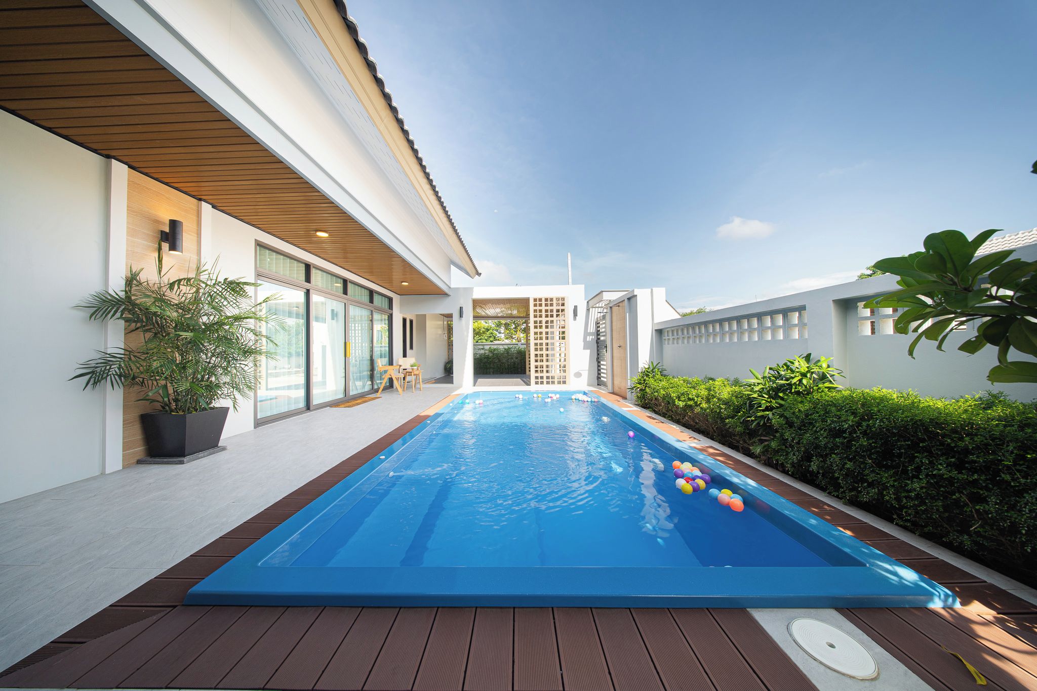 Une piscine avec une terrasse en bois, entourée d’une maison moderne dotée de grandes fenêtres et d’un jardin.