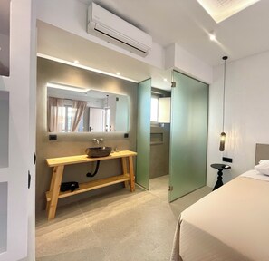 Ein modernes Hotelzimmer mit Schminktisch aus Holz, einem großen Spiegel, einer eingebauten Klimaanlage, einem Bett mit weißer Bettwäsche und einem kleinen runden Tisch mit Lampe.
