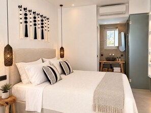 A bedroom with a bed, hanging tassels, pendant lights, a bedside table with a plant, and a view of a bathroom. - Mykonos Dot Suites (Ano Mera)