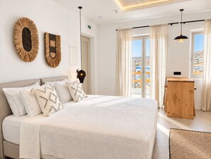 A bedroom with a large bed, a wooden side table, and a view of the city through the window. - Mykonos Dot Suites (Ano Mera)