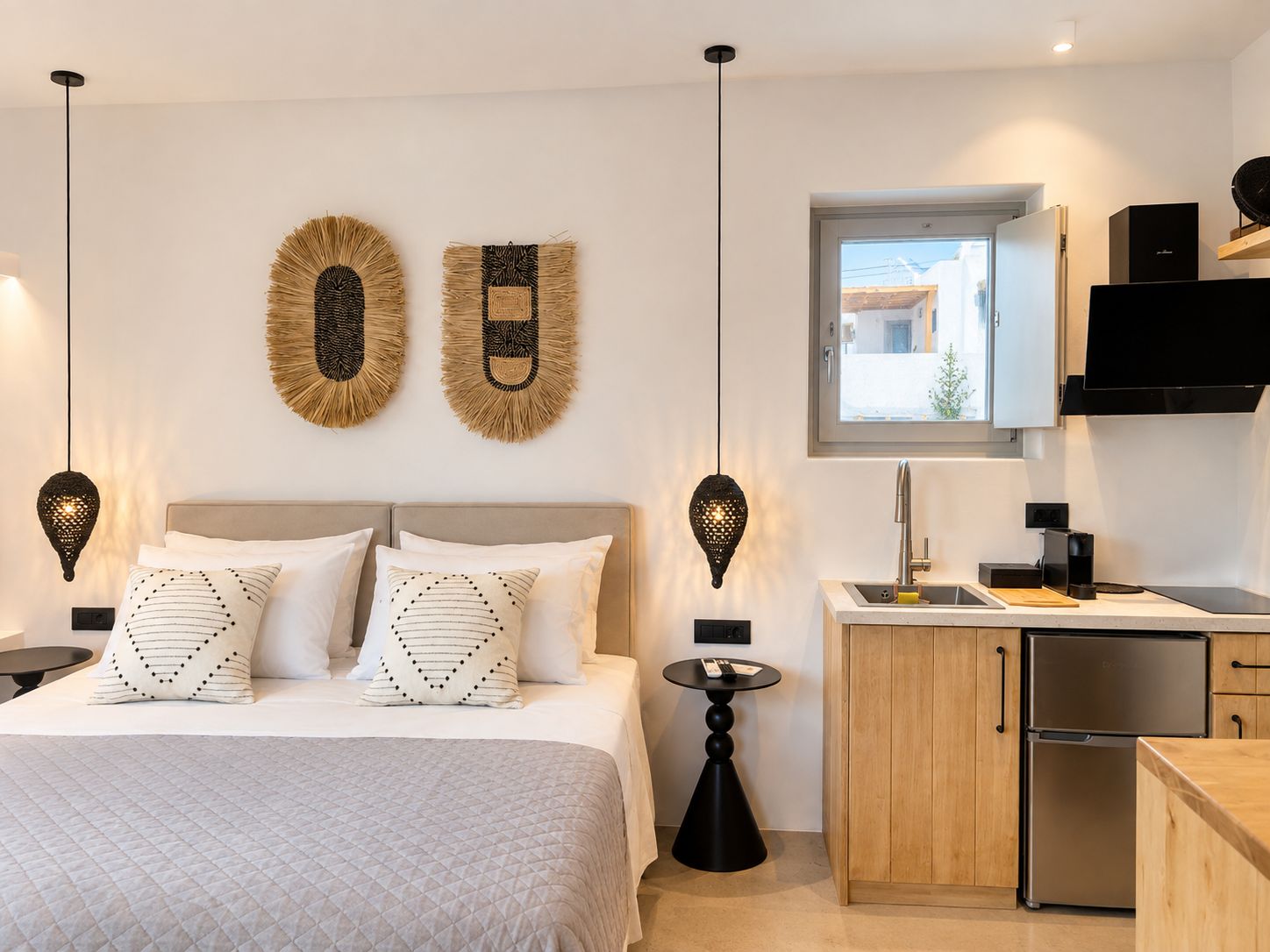 Ein modernes Schlafzimmer mit einem Bett, Nachttischen und einem kleinen Tisch daneben. Es gibt eine Küchenzeile mit Spüle, Herd und Kühlschrank.