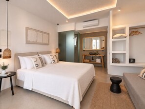 A modern bedroom with a large bed, bedside tables, a vanity with a mirror, and a seating area with a sofa and a small table. - Mykonos Dot Suites (Ano Mera)