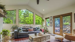 A spacious living room with a large gray sectional sofa, a coffee table, and a patterned rug. There are several potted plants and a large window providing a view of trees. - Hot tub, Outdoor dining, BBQ Grill, Fire pit & Gym (Colorado Springs)
