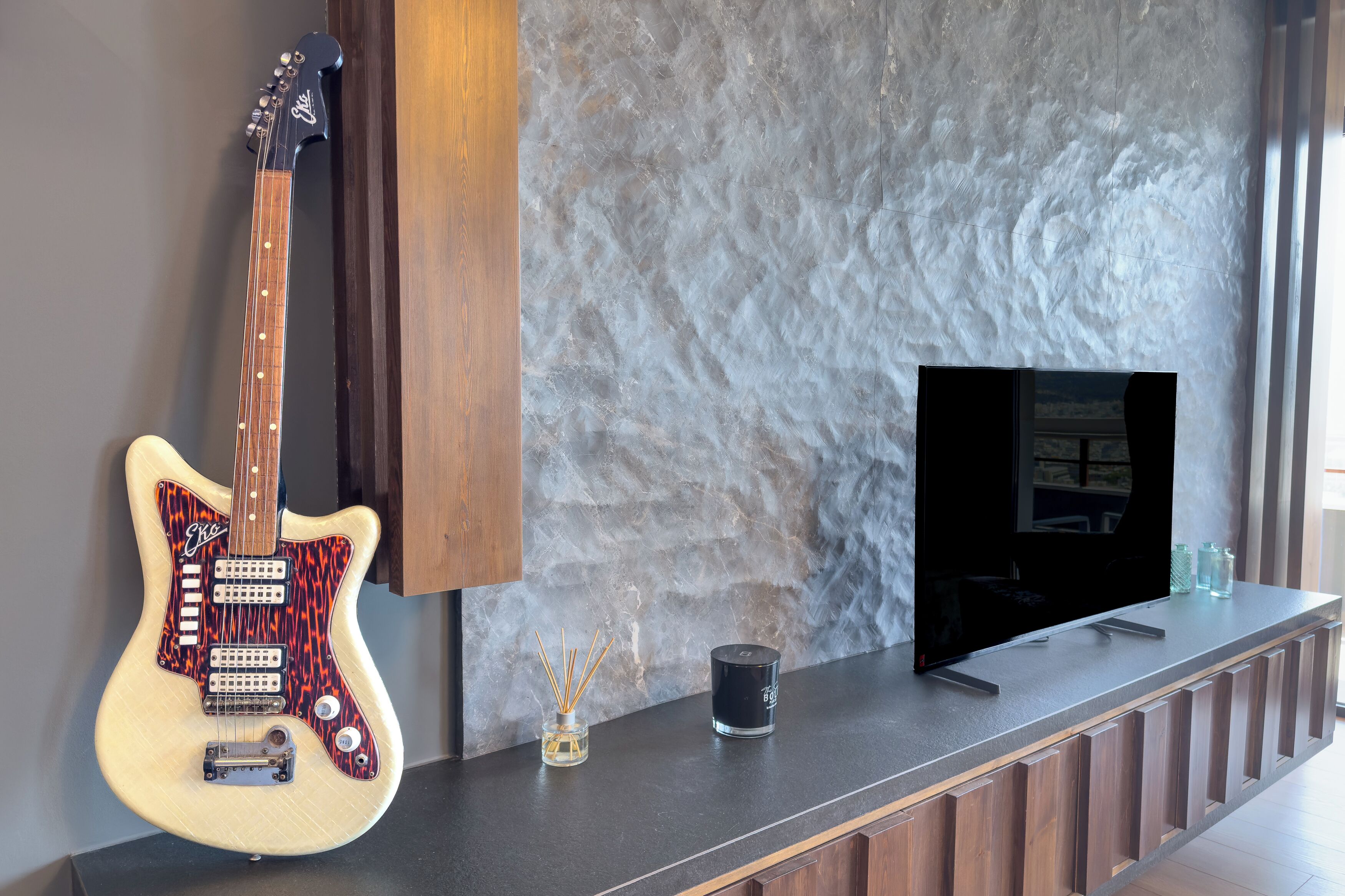 A modern room with a marble wall, a wooden cabinet, a television, and a guitar.