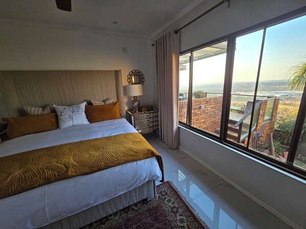 Une chambre à coucher avec un grand lit, une table de chevet, une lampe et une vue sur la ville grâce à de grandes fenêtres.