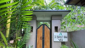 A gated entrance with a wooden door, surrounded by lush greenery. A sign reads "D'Bunut Villa."