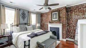 A bedroom with a brick wall, a fireplace, a bed with a headboard, a bench, and a ceiling fan.