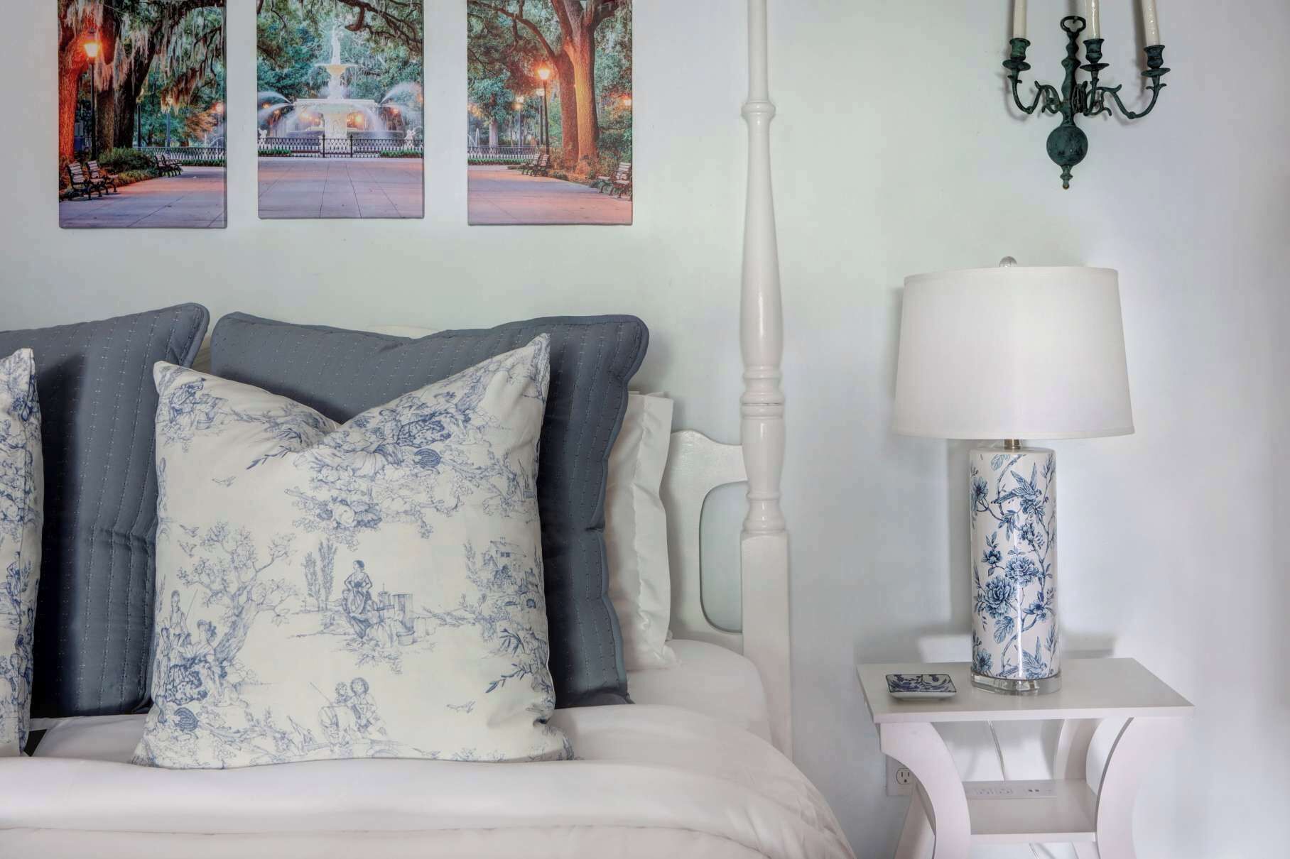 A neatly made bed with a white headboard, grey and blue patterned pillows, a white bedside table with a lamp, and a wall-mounted chandelier.