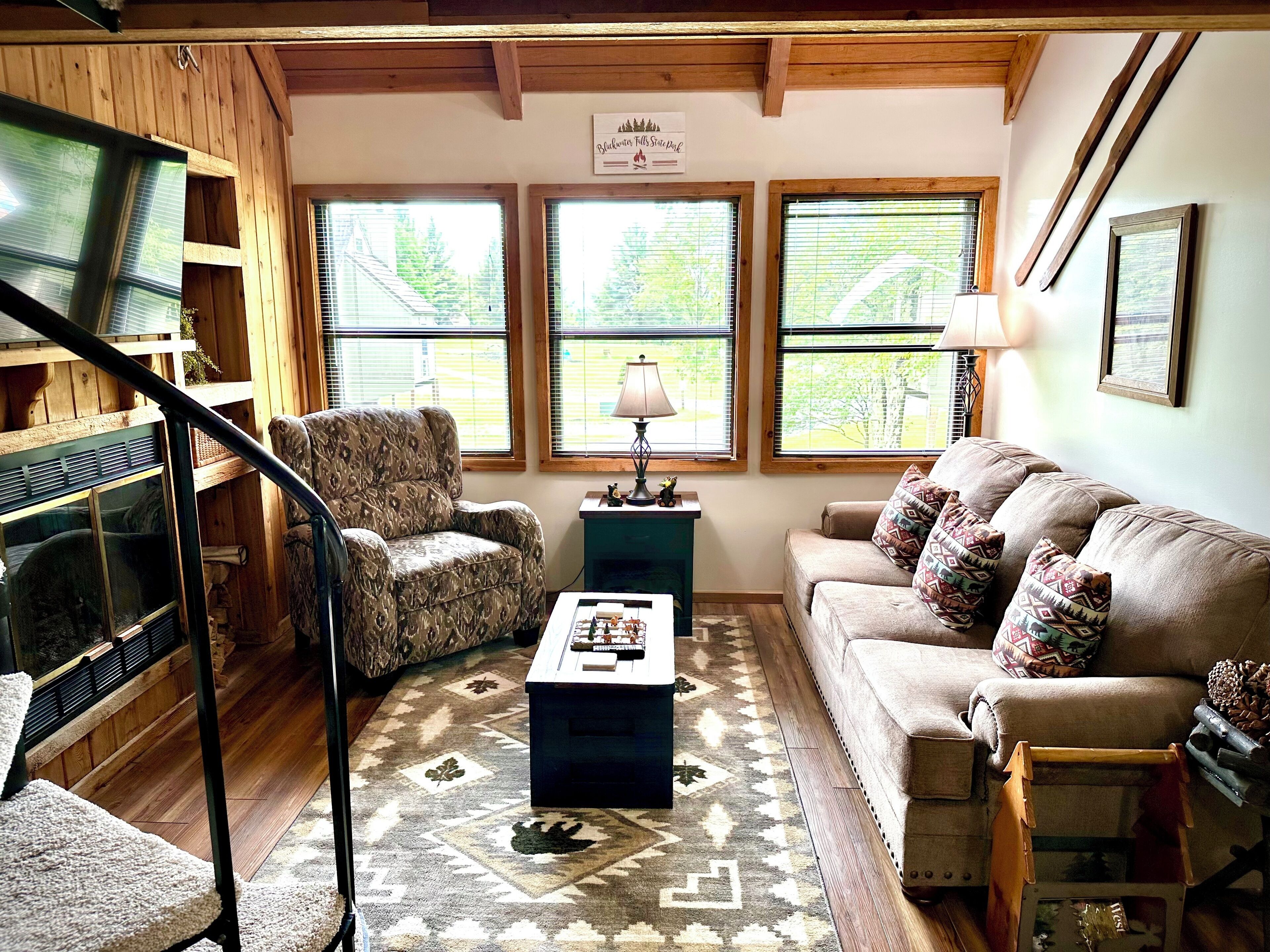 A cozy living room with a wooden ceiling, a patterned armchair, a beige sofa with decorative pillows, a coffee table, and a large window with a view of trees.