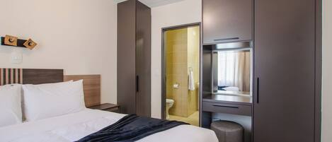 Habitación de hotel moderna con una cama grande, una mesita de noche, un armario y un baño visible a través de una puerta de vidrio.