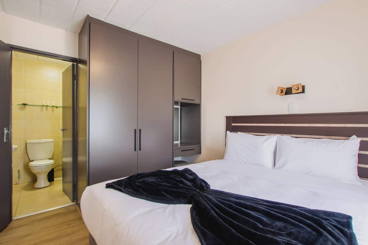 Quarto de hotel com cama, guarda-roupa, televisão e banheiro com vaso sanitário e chuveiro.