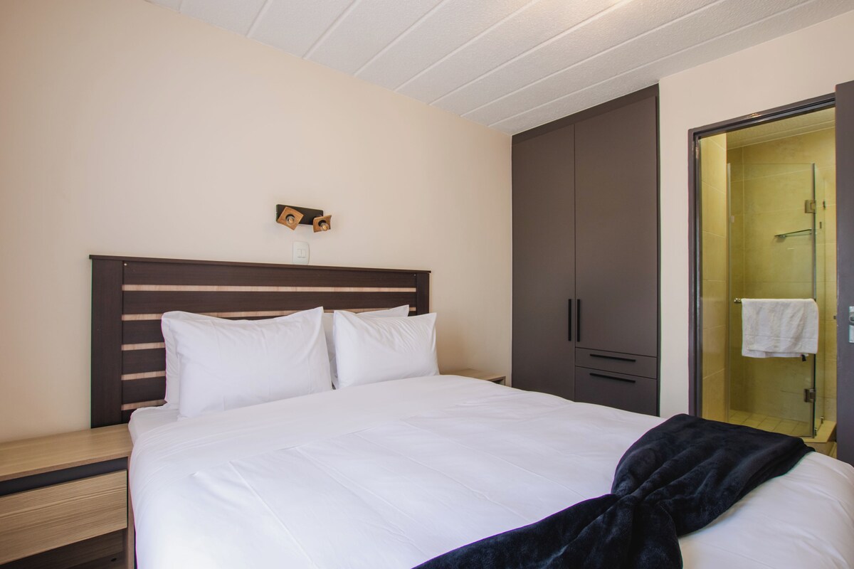 Ein Hotelzimmer mit einem großen Bett, einem Holzkopfende, einem Nachttisch, einem Kleiderschrank und einer Duschkabine aus Glas.