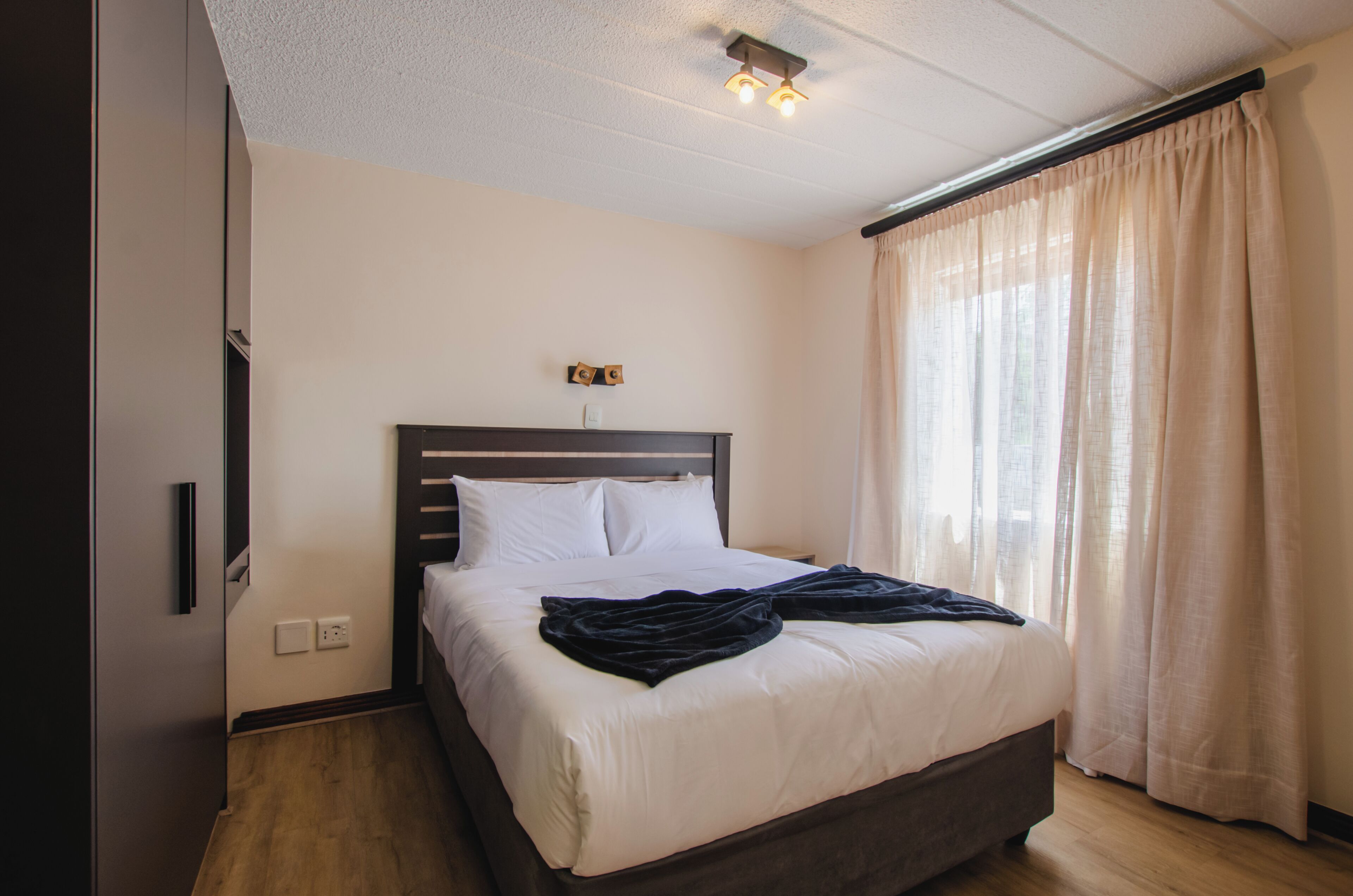 Een net opgemaakt bed met witte lakens en een donker dekbed, een houten hoofdbord, en een raam met beige gordijnen.