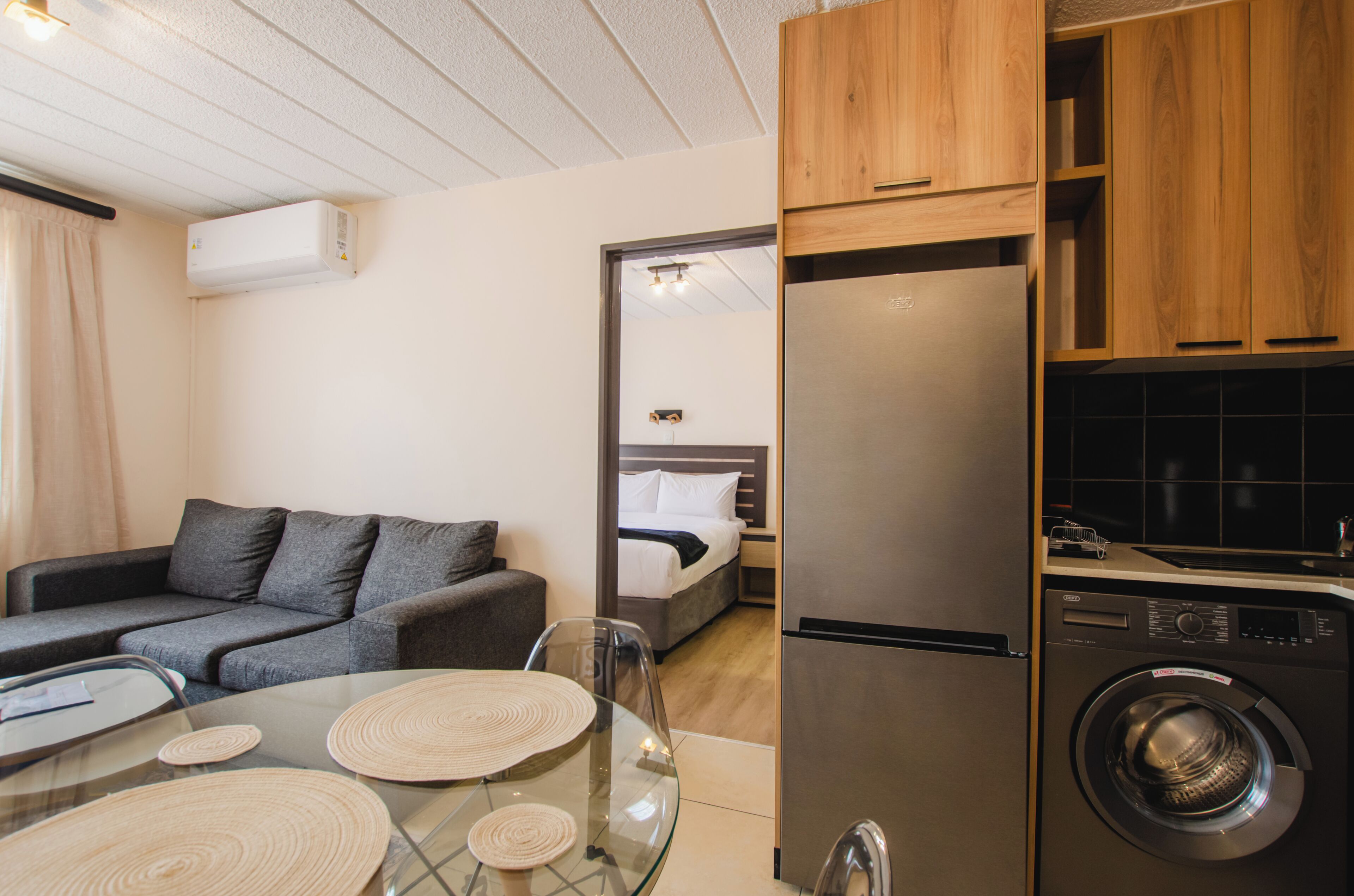Un espace de vie compact comprenant un canapé gris, une table à manger en verre, un lave-linge et un coin cuisine de taille réduite.