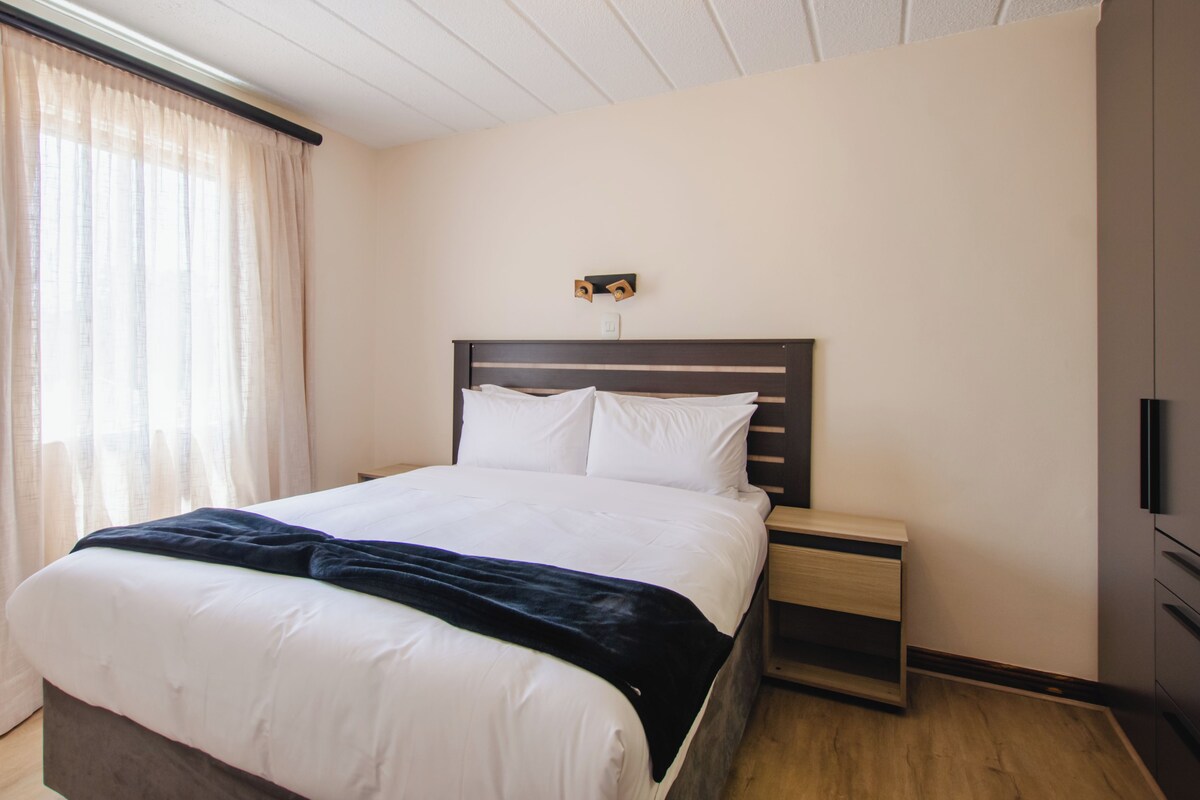A neatly made bed with white linens and a dark blanket, a wooden nightstand with a drawer, and a wall-mounted light fixture.