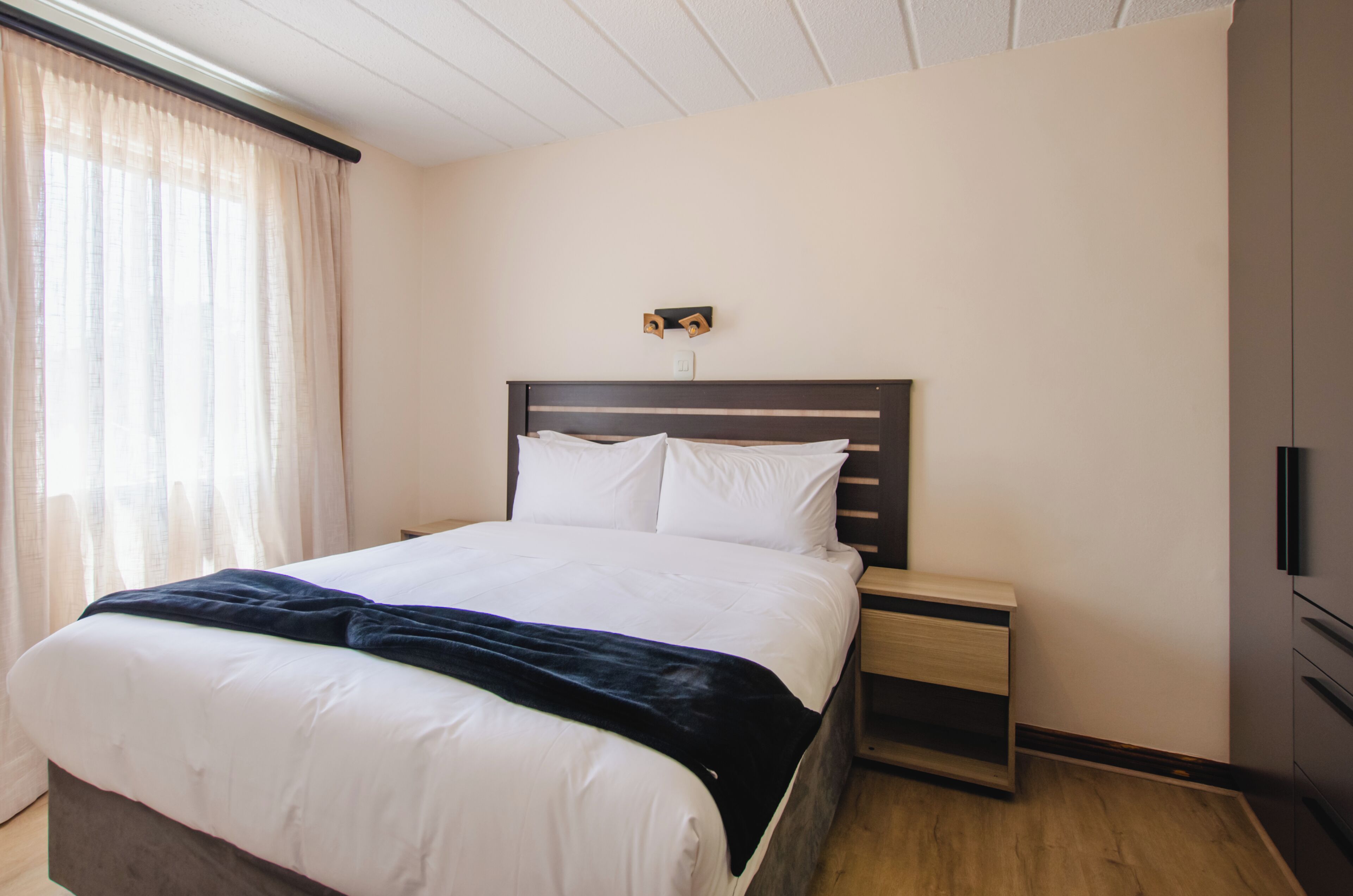 A neatly made bed with white linens and a dark blanket, a wooden nightstand with a drawer, and a wall-mounted light fixture.