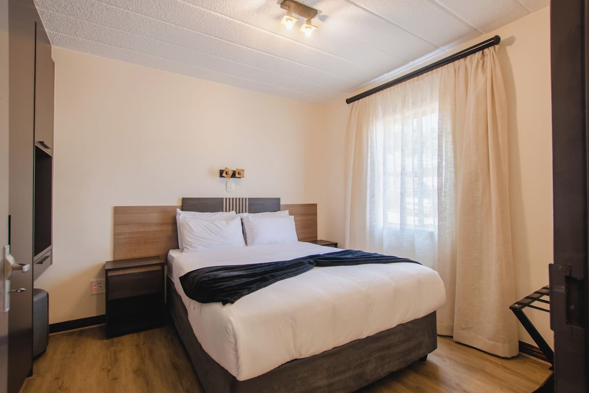 Quarto de hotel com cama, mesa de cabeceira, televisão e janela com cortinas.