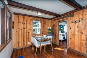 A wooden-paneled room with a dining table set for four, a window, and a kitchen area visible through an open doorway.
