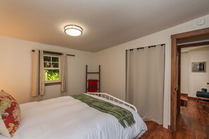Un dormitorio con cama, ventana con cortinas, escalera de madera y puerta que da a otra habitación.