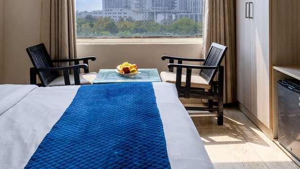 A hotel room with a large window, two chairs, a bed with a blue quilt, and a small table with fruit. - Nexhub Palace (Gurugram)