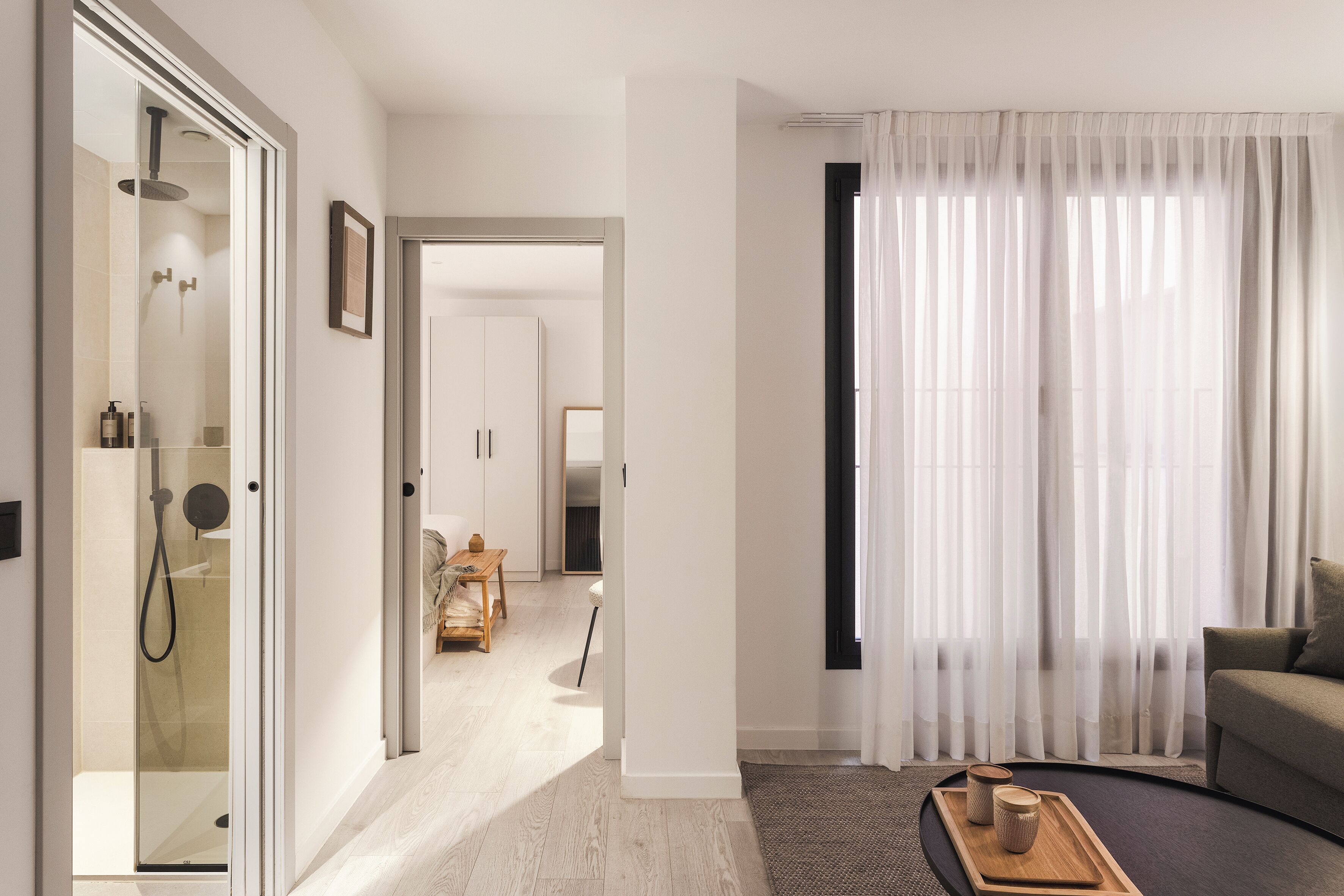Une chambre d’hôtel moderne dotée d’une douche à parois de verre, d’un canapé, d’un coin repas et d’une grande fenêtre avec des rideaux légers.