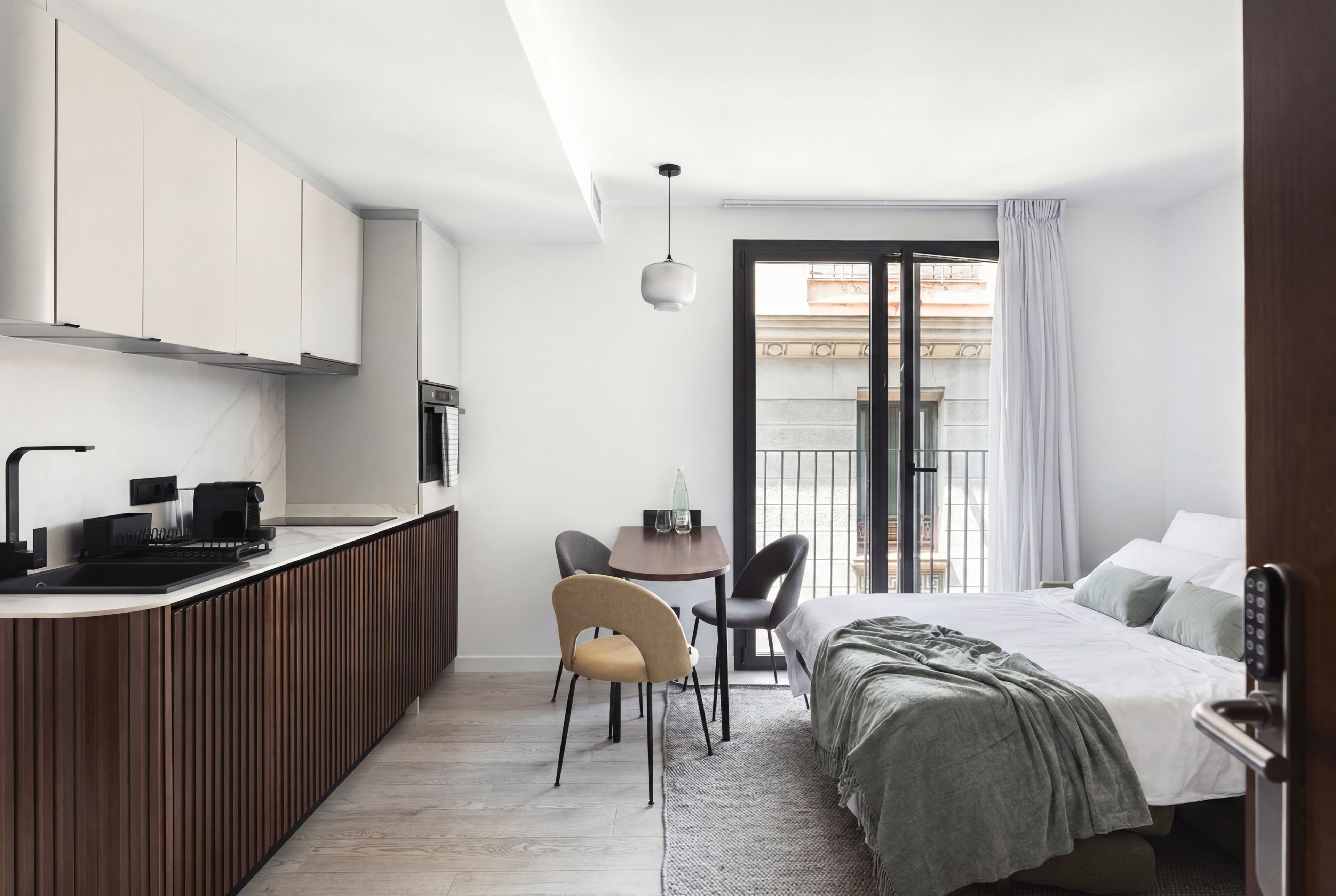 Habitación de hotel pequeña con kitchenette, una cama con manta verde, una mesa de comedor con sillas y vistas a través de un ventanal.