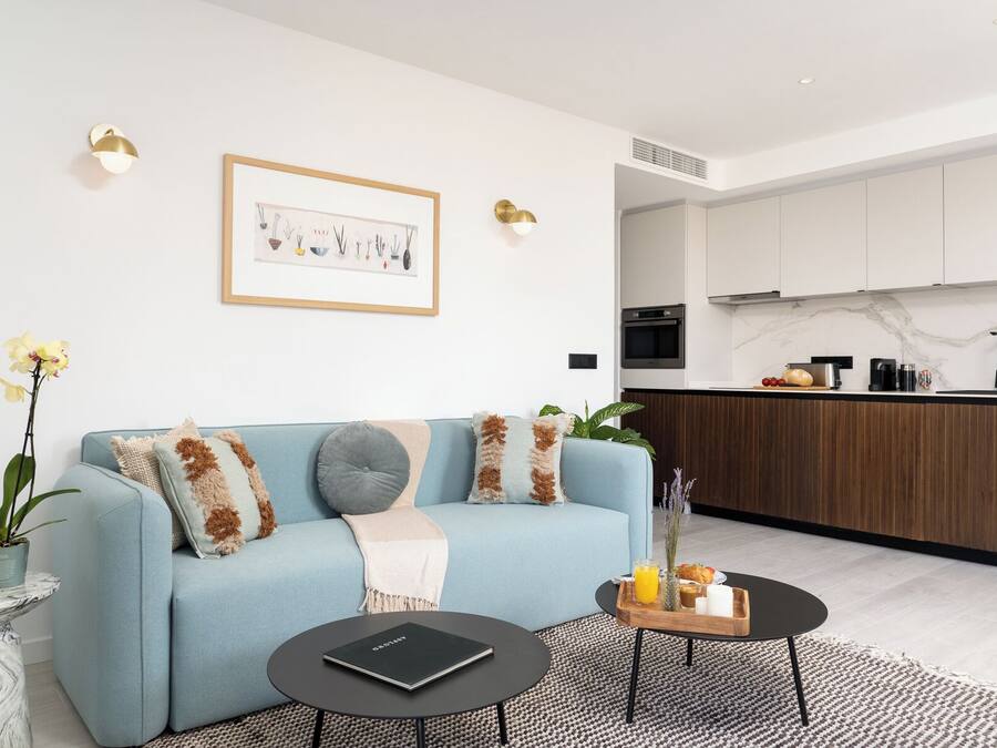A modern living room with a light blue sofa, a coffee table, and a small side table with a book. A kitchen area with white cabinets and a marble countertop is visible in the background.
