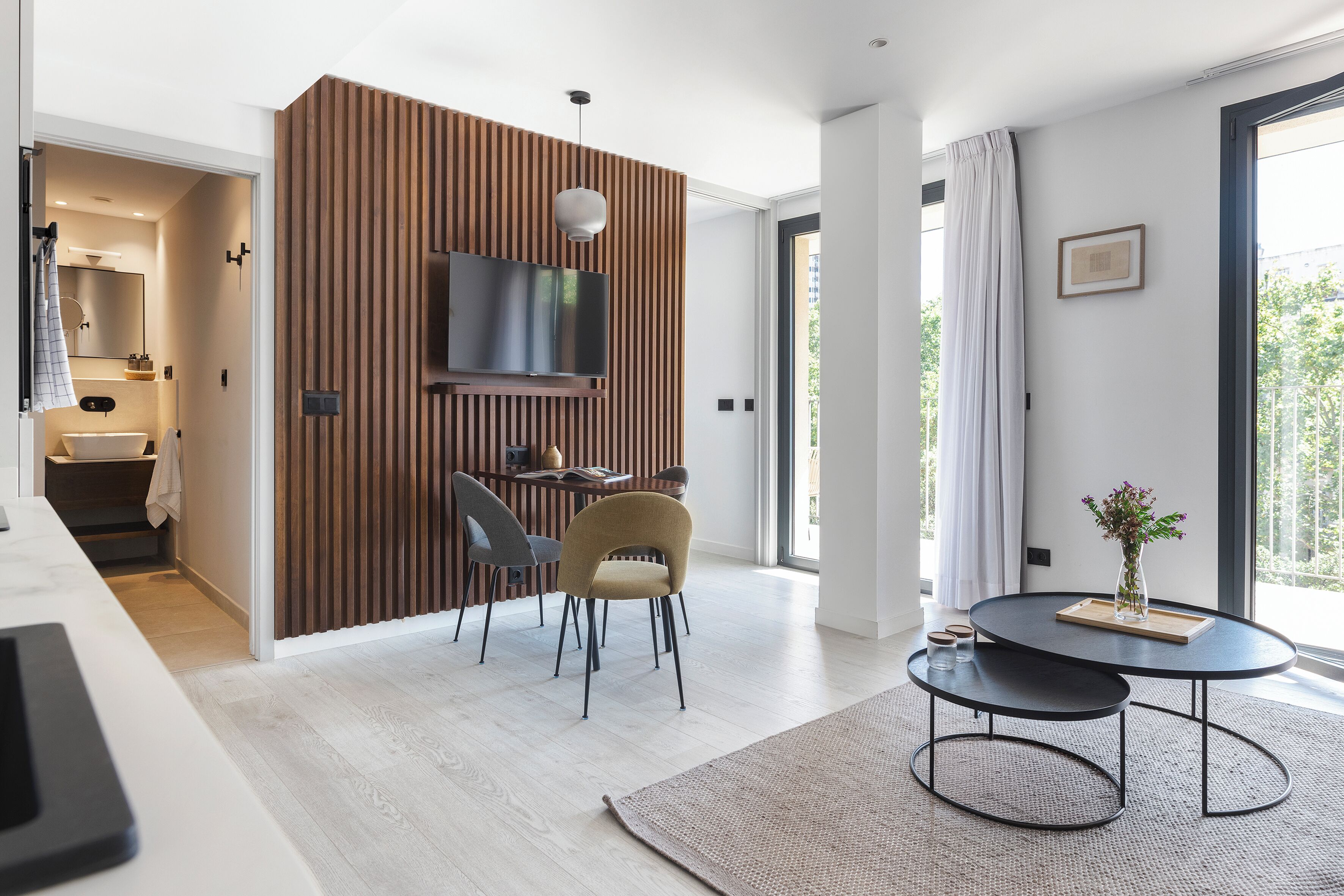 Una sala de estar moderna con televisión, un comedor con sillas, una mesa de centro redonda y vistas al exterior.