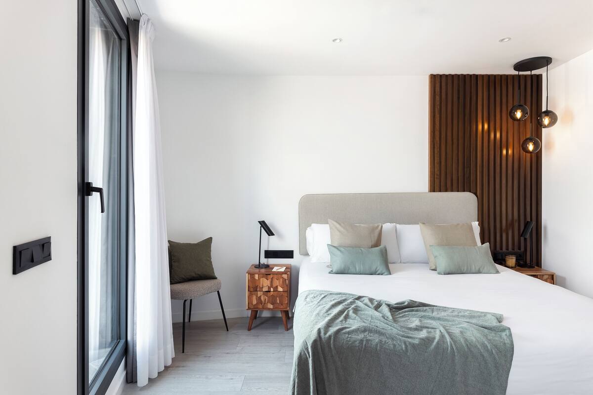 Quarto moderno com uma cama grande, mesa de cabeceira de madeira, uma cadeira e uma janela com cortinas.