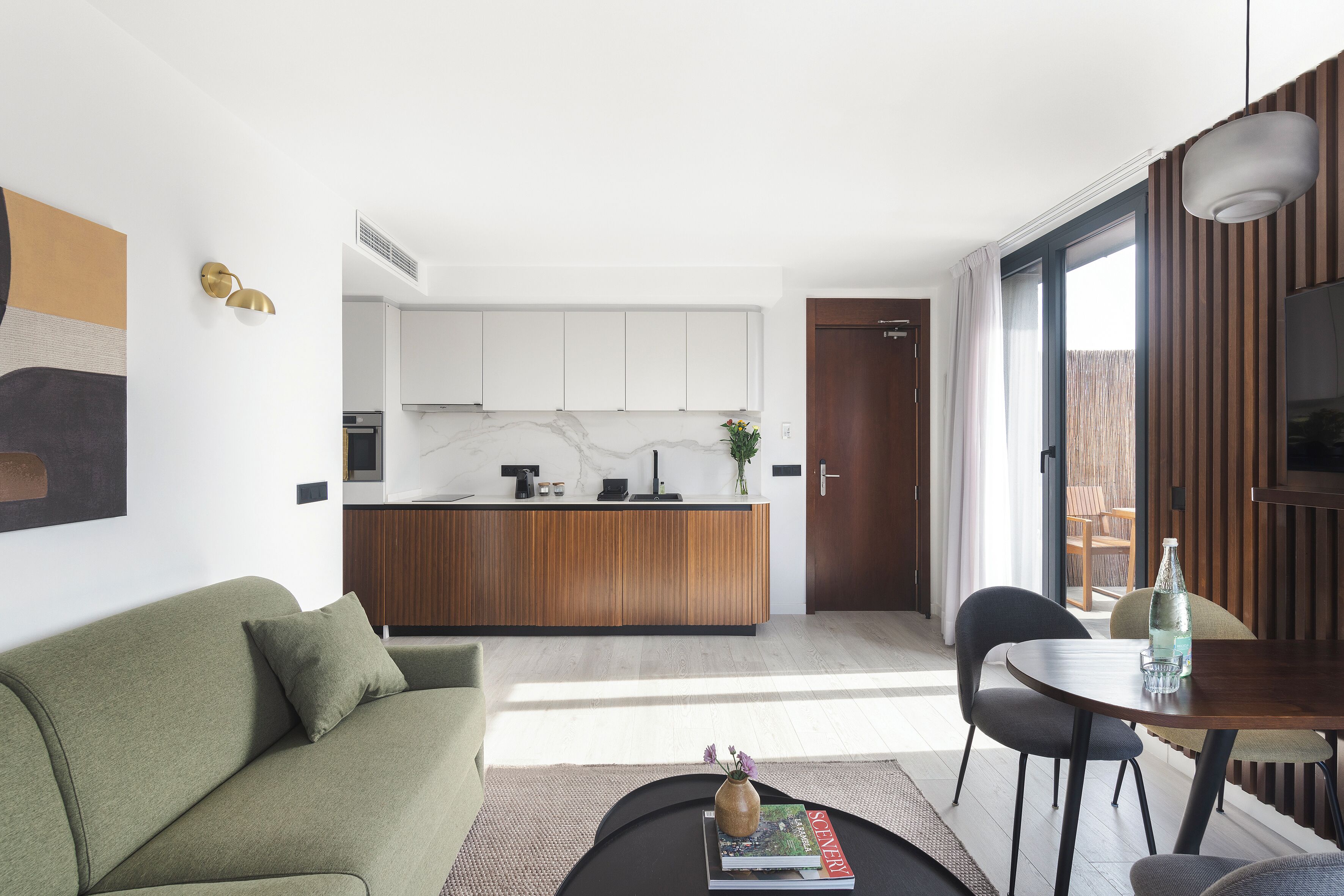 A modern living space with a sofa, a coffee table with a book and vase, a dining area with chairs, a kitchen with white cabinets, and a sliding glass door to an outdoor area.