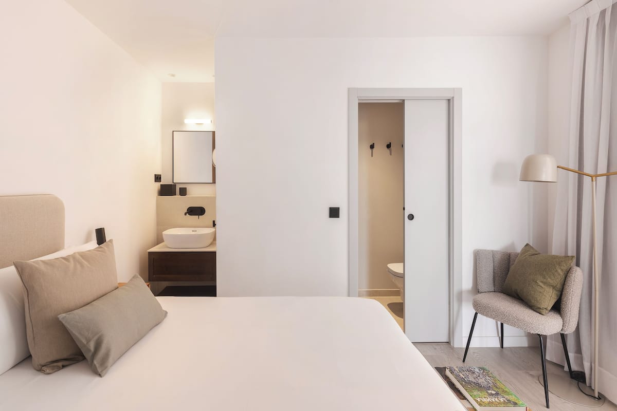 Quarto de hotel moderno com cama grande, mesa de cabeceira, cadeira, espelho e banheiro visível através de uma porta aberta.