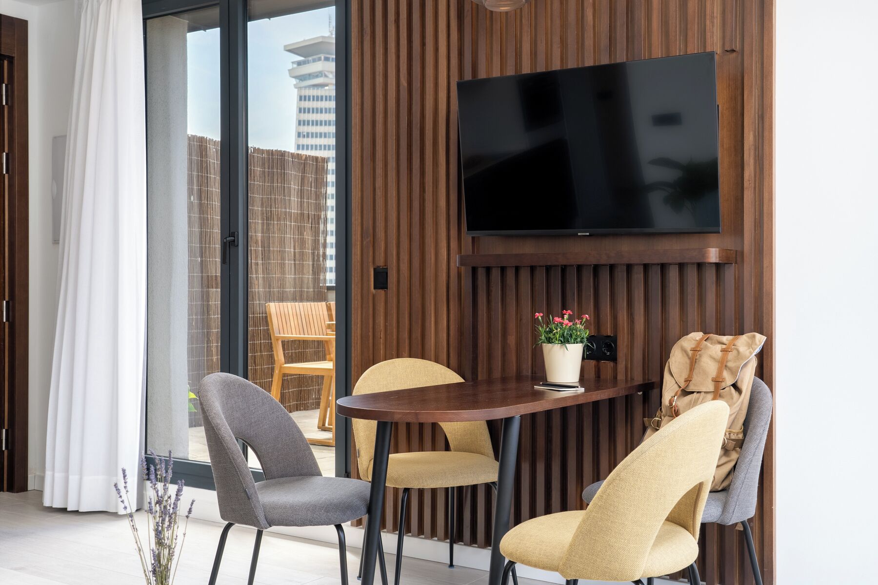 Un salón moderno con una mesa redonda de madera, dos sillas grises y dos amarillas, un televisor de pantalla plana montado en la pared y una planta en maceta sobre la mesa.