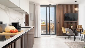Une cuisine moderne avec un plan de travail en marbre, une grande planche à découper sur laquelle on voit des tomates, et une machine à café. Il y a également un coin repas avec une table, des chaises et un téléviseur fixé au mur.