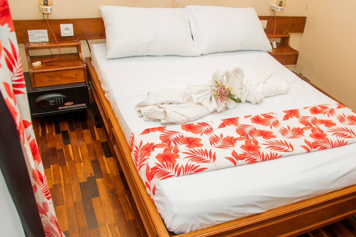 Un lit bien fait, recouvert d’une couvre-lit à motifs floraux, avec un oreiller blanc et une serviette ornée de fleurs. On y trouve une table de chevet en bois avec un téléphone et une petite table avec une lampe. La pièce est dotée d’un plancher en bois et d’une tête de lit en bois.