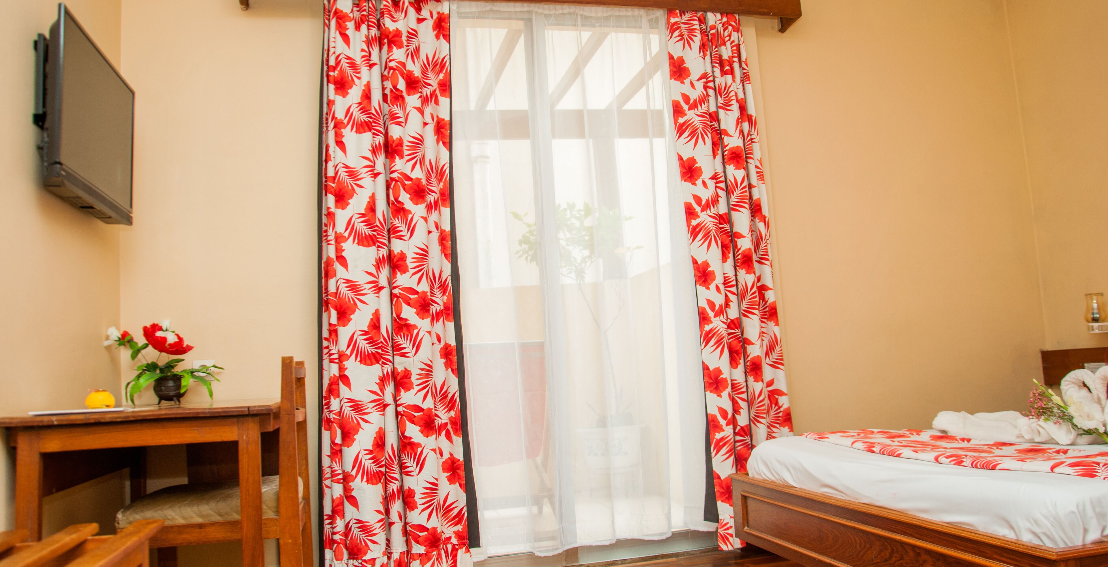 A hotel room with a bed, a wooden desk, a chair, a TV mounted on the wall, and a window with red and white floral curtains.