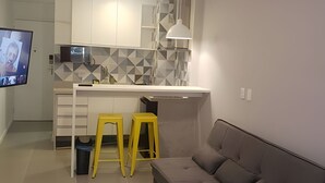 A modern kitchen with a white countertop, two yellow stools, a grey sofa, and a flat-screen TV. - Studio Copacabana a 100 metros da Praia (Rio de Janeiro)