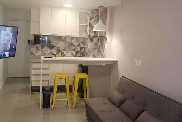 Una cocina moderna con encimera blanca, dos taburetes amarillos, un sofá gris y un televisor de pantalla plana.
