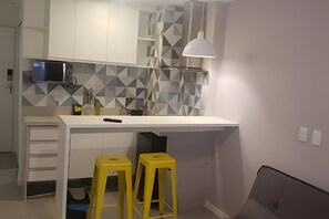 Cocina compacta con encimera blanca, dos taburetes amarillos, lámpara colgante moderna y salpicadero con estampado geométrico.