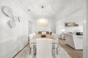 A spacious dining area with a wooden table and white upholstered chairs, a hanging light fixture, and a white wall with abstract art.