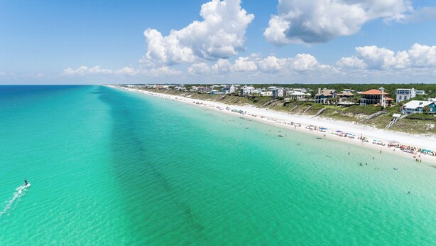 A coastal beach with clear turquoise waters, a sandy shore, and beachfront properties.