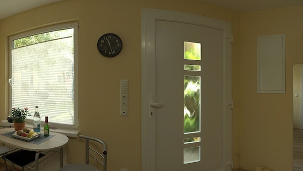 A room with a dining table, chairs, a clock, a door with frosted glass panels, and a window with blinds. - Small apartment for two people with garden and private entrance (Bochum)