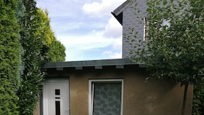 A small, single-story building with a door and a window, surrounded by a well-maintained lawn and a dense hedge. - Small apartment for two people with garden and private entrance (Bochum)