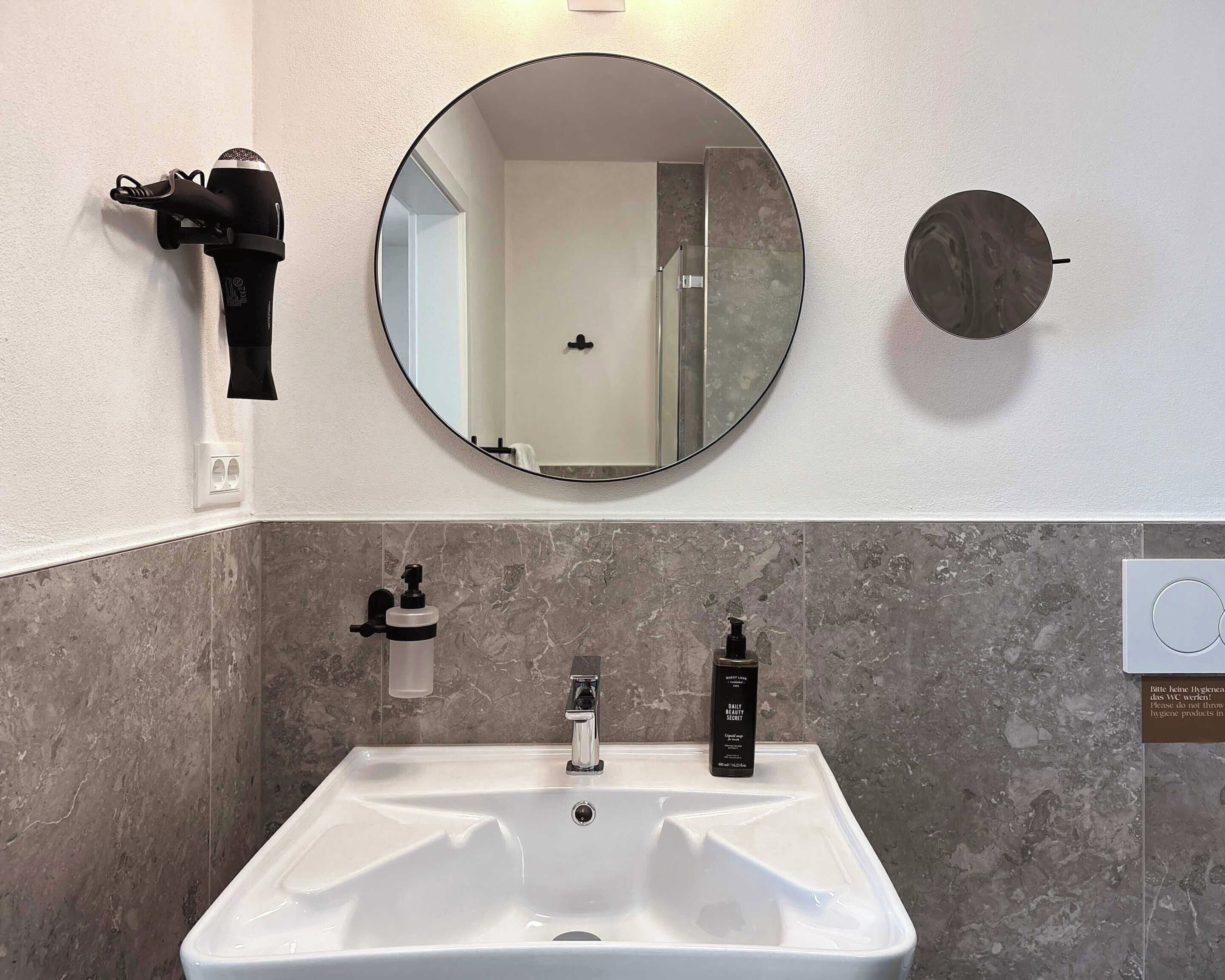 A bathroom with a round mirror, a hairdryer, a sink with a faucet, and a bottle of hand soap.