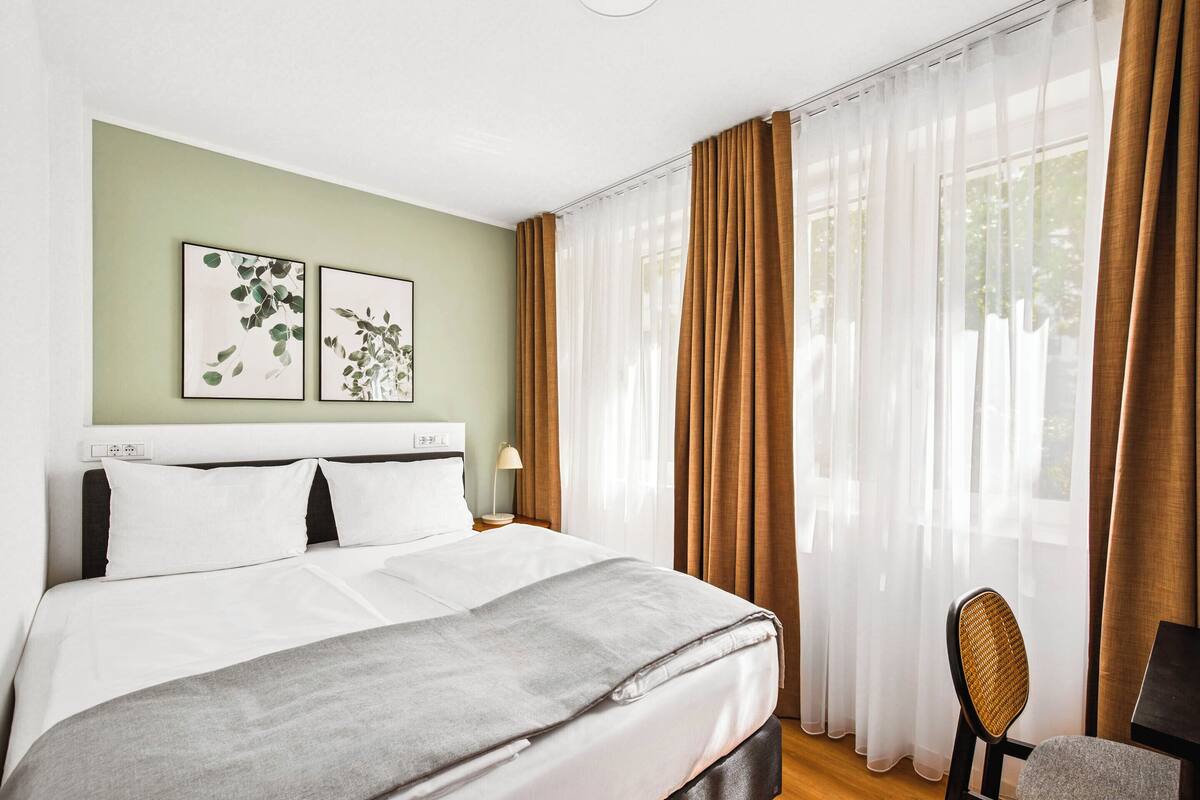 A neatly made bed with white linens, a grey throw blanket, and a headboard with a white panel. There are two framed artworks on the wall above the bed, and a chair is visible to the right.