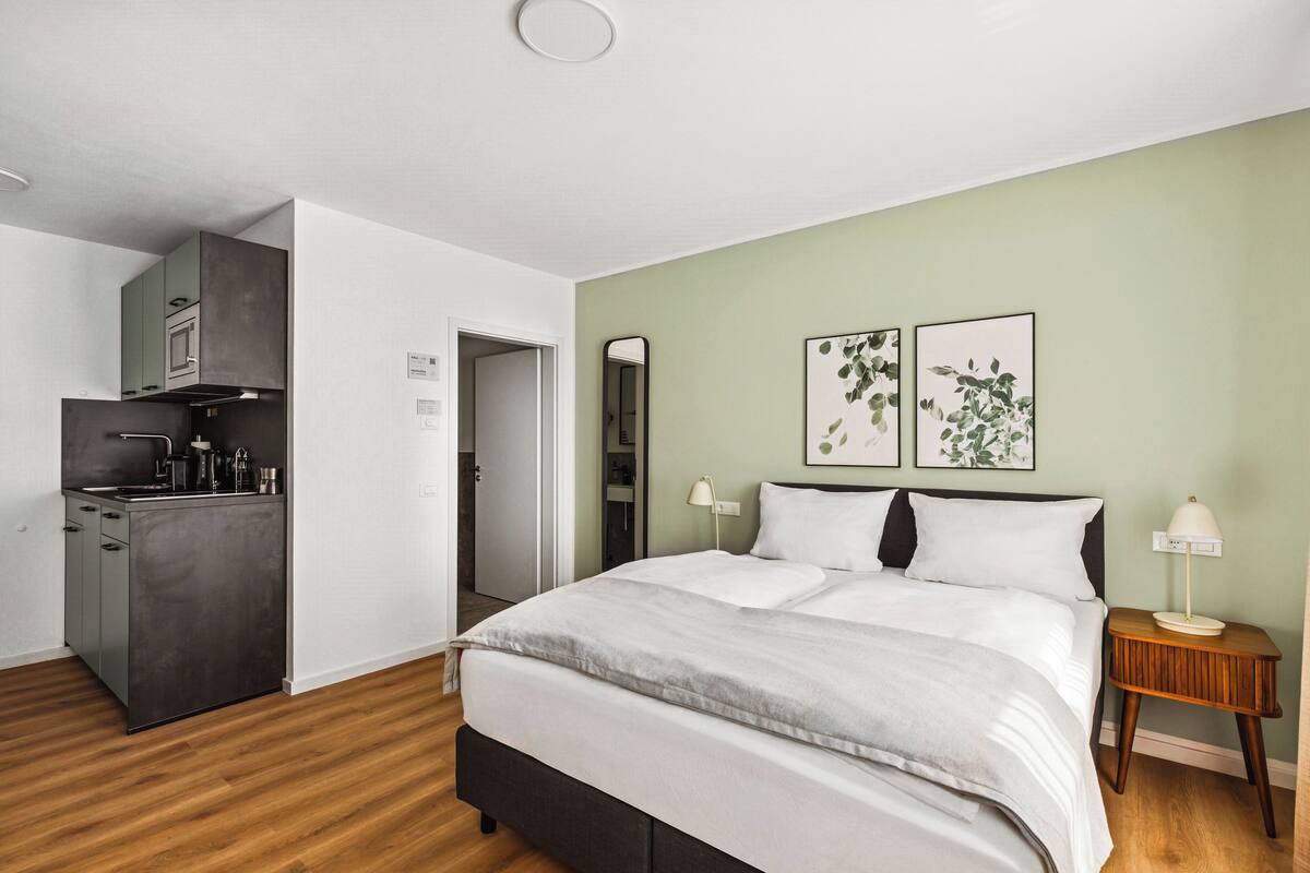 A modern bedroom with a large bed, a bedside table with a lamp, and a view of a kitchen area.