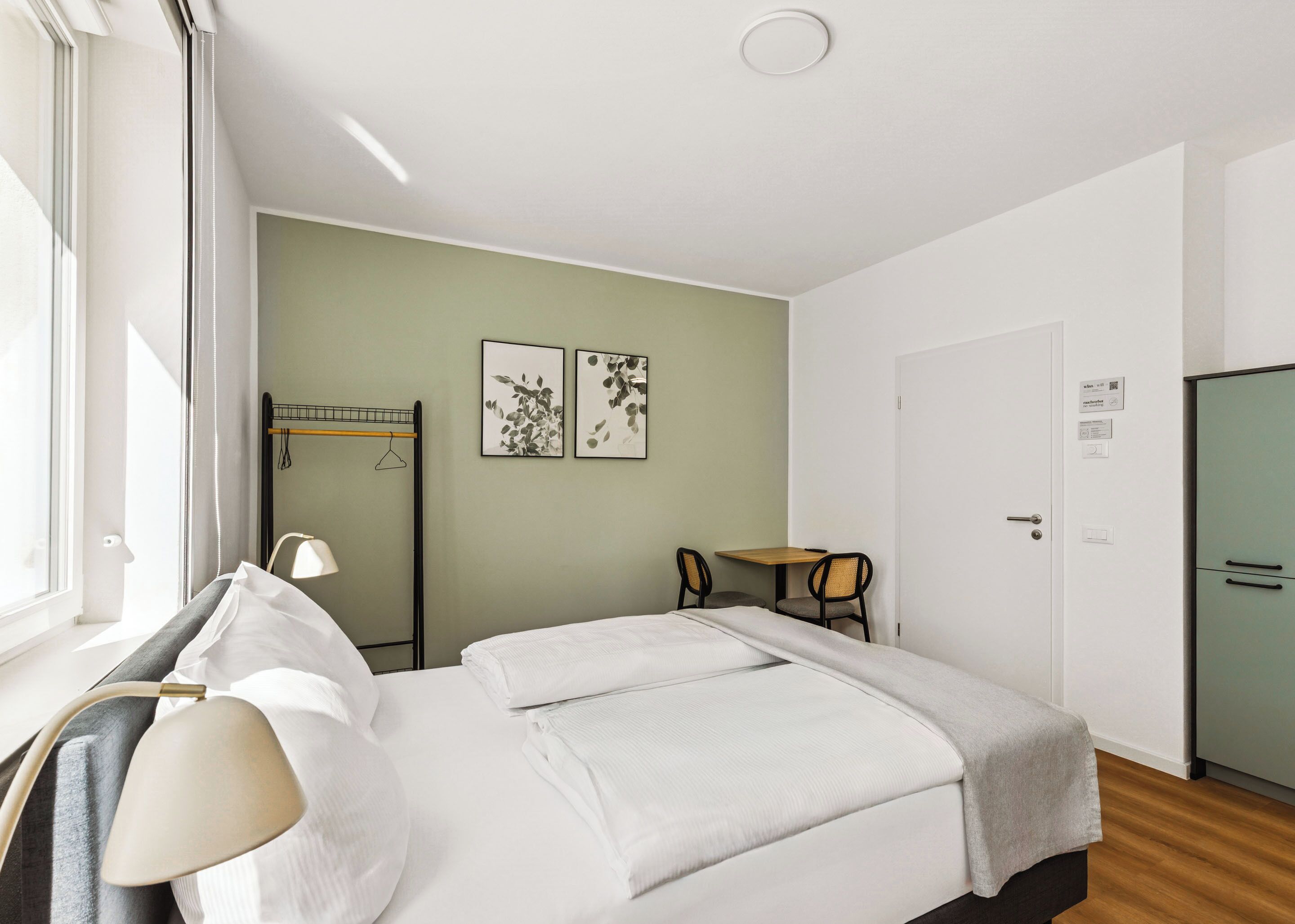 Een netjes ingerichte hotelkamer met een bed, nachtkastje, lamp en een kleine tafel met stoelen.