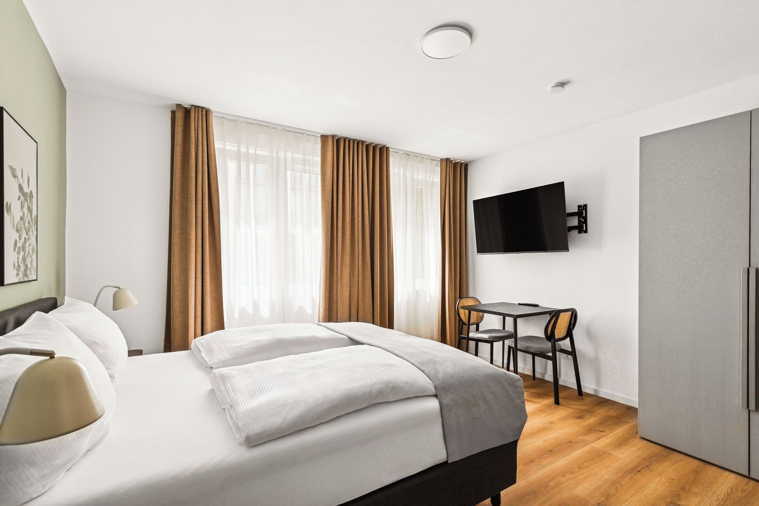 Ein modernes Hotelzimmer mit einem großen Bett, einem Schreibtisch mit Stuhl, einem an der Wand montierten Fernseher und einem Kleiderschrank.