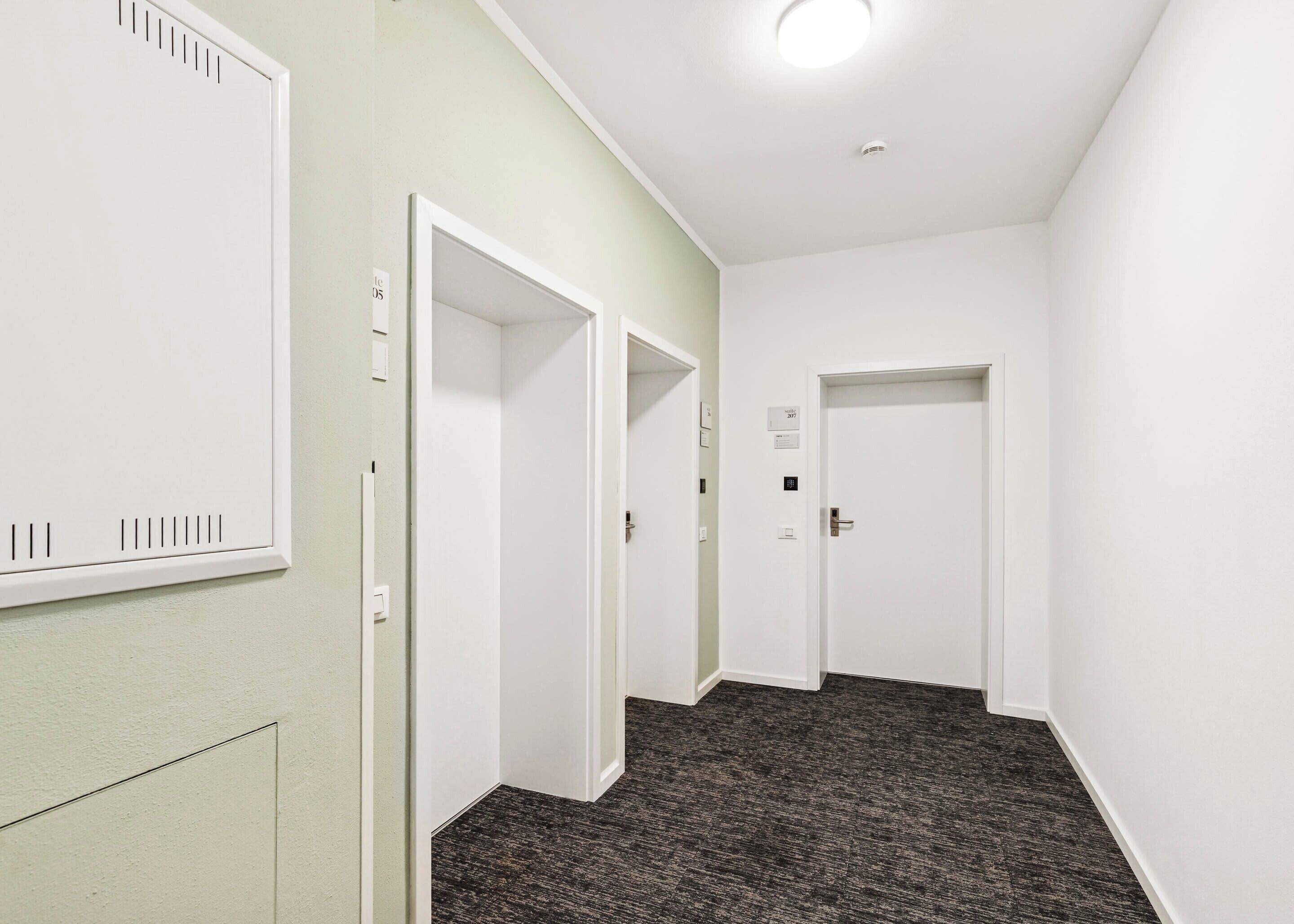 A hallway with white walls, a carpeted floor, and doors leading to individual rooms.