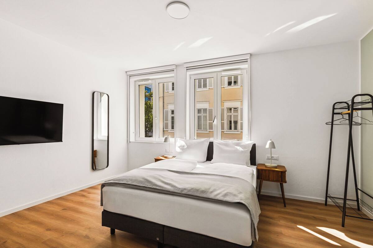 A modern bedroom with a bed, a TV mounted on the wall, a bedside table with a lamp, and a window with a view of buildings.